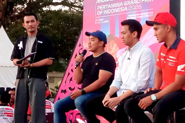 Four men sit on a stage at an outdoor event; one man in a black shirt holds a microphone while others listen, with a bright pink promotional backdrop behind them.