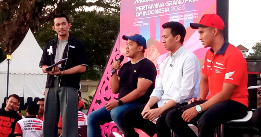 Four men sit on a stage at an outdoor event; one man in a black shirt holds a microphone while others listen, with a bright pink promotional backdrop behind them.