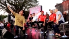 Man in a tan shirt takes a selfie on a stage with panelists seated behind him in red shirts at an outdoor event.