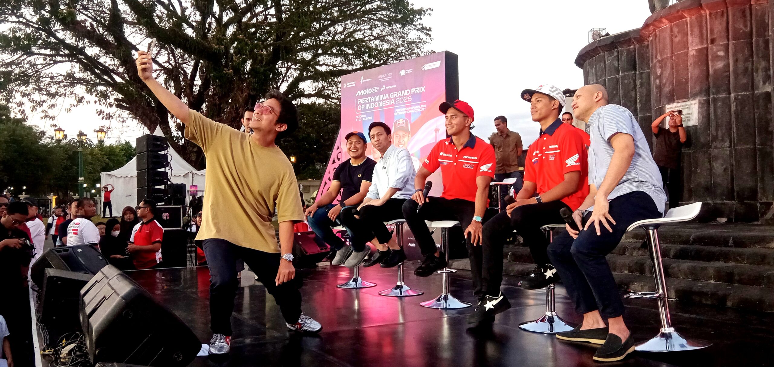 Man in a tan shirt takes a selfie on a stage with panelists seated behind him in red shirts at an outdoor event.