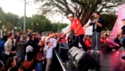 Staff on stage interacting with a crowd at an outdoor event as a man in red hands a cap to a child in front of the audience, with a speaker and banner in the background.