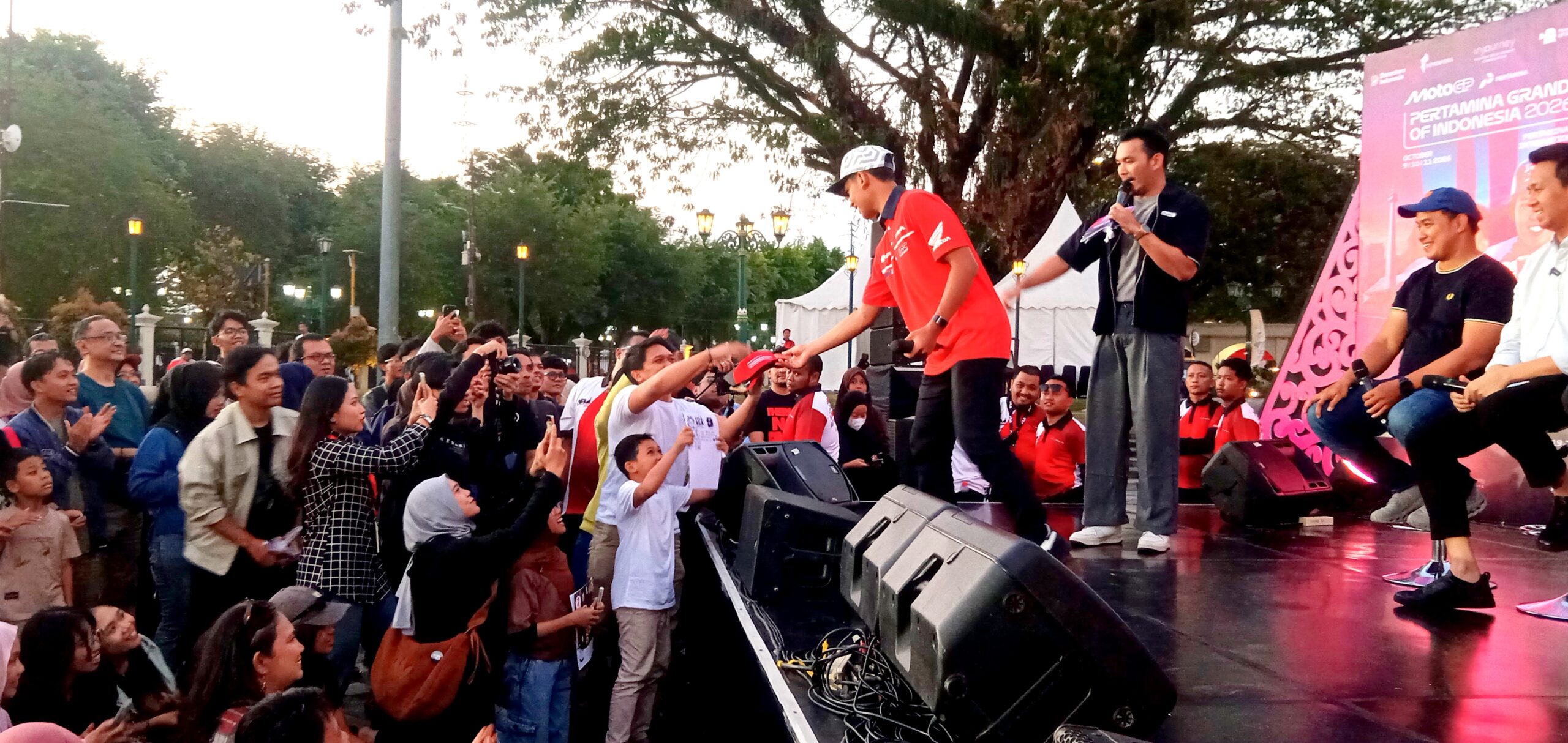 Staff on stage interacting with a crowd at an outdoor event as a man in red hands a cap to a child in front of the audience, with a speaker and banner in the background.