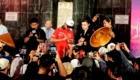 Crowd gathers at a stage as a man in a red racing outfit signs autographs while photographers snap photos nearby.