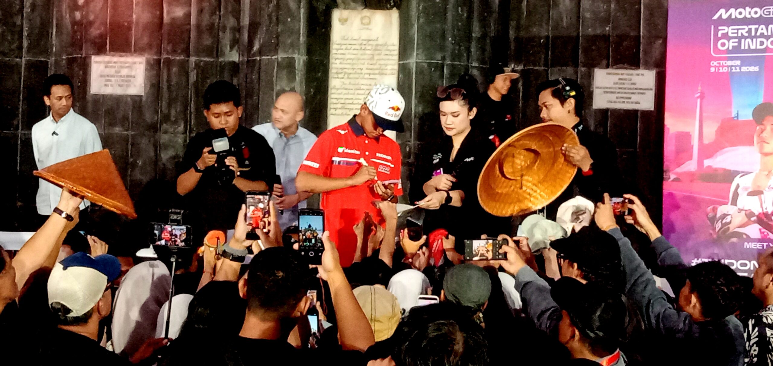 Crowd gathers at a stage as a man in a red racing outfit signs autographs while photographers snap photos nearby.