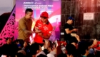 Rider in red racing jersey signs autographs for fans as a man in a brown shirt assists, on a stage with a purple backdrop reading Pertamina Grand Prix of Indonesia 2026 and a crowd watching.