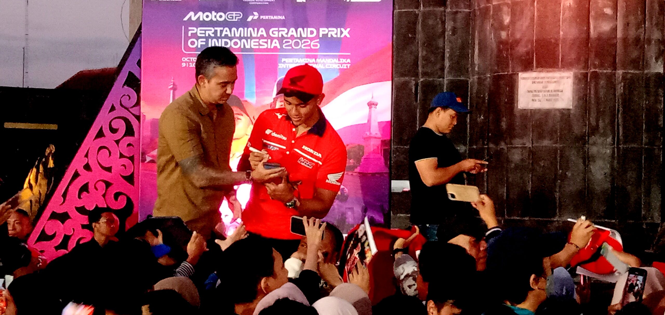 Rider in red racing jersey signs autographs for fans as a man in a brown shirt assists, on a stage with a purple backdrop reading Pertamina Grand Prix of Indonesia 2026 and a crowd watching.