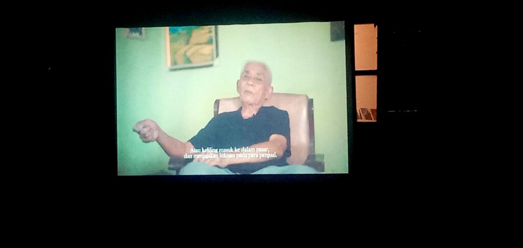 Man in a dark shirt sits in a beige chair, displayed on a green wall video with Indonesian subtitles at the bottom