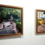 Two framed paintings on a white gallery wall; left image shows a forest path with people walking, right image shows a rural dirt road with a wagon drawn by oxen.