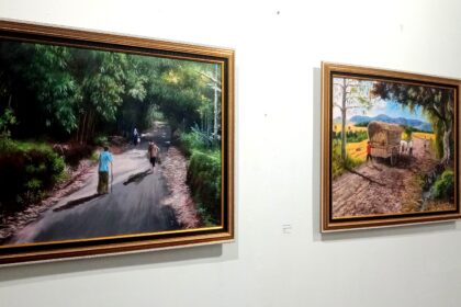 Two framed paintings on a white gallery wall; left image shows a forest path with people walking, right image shows a rural dirt road with a wagon drawn by oxen.