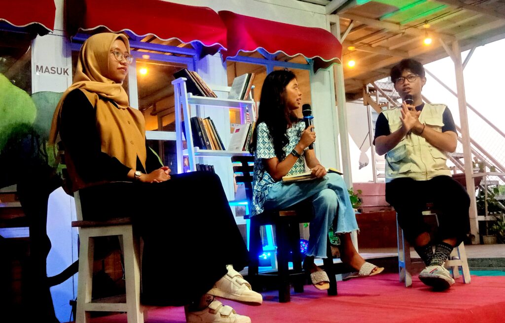 Panel discussion on stage with three people: woman in hijab on left, woman with microphone in center, man with microphone on right; library shelves and string lights in background.