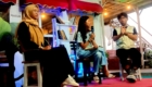 Panel discussion on stage with three people: woman in hijab on left, woman with microphone in center, man with microphone on right; library shelves and string lights in background.