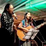 Two women perform on a small stage: one singing into a microphone, the other playing an acoustic guitar with a second mic nearby.