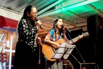 Two women perform on a small stage: one singing into a microphone, the other playing an acoustic guitar with a second mic nearby.
