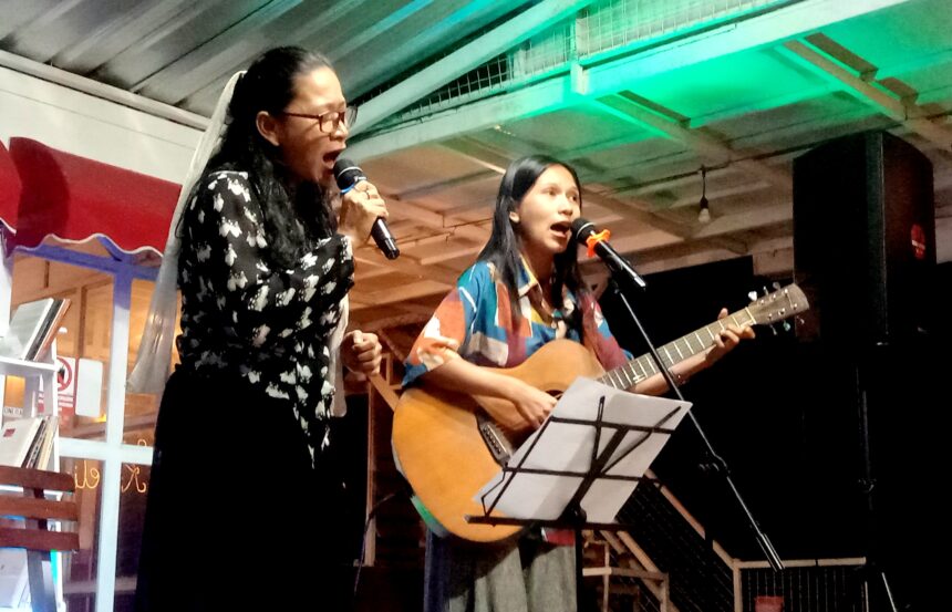 Two women perform on a small stage: one singing into a microphone, the other playing an acoustic guitar with a second mic nearby.