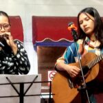 Two women perform on a small stage: left harmonica player with glasses, right singer-guitarist at a microphone.