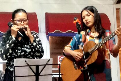 Two women perform on a small stage: left harmonica player with glasses, right singer-guitarist at a microphone.