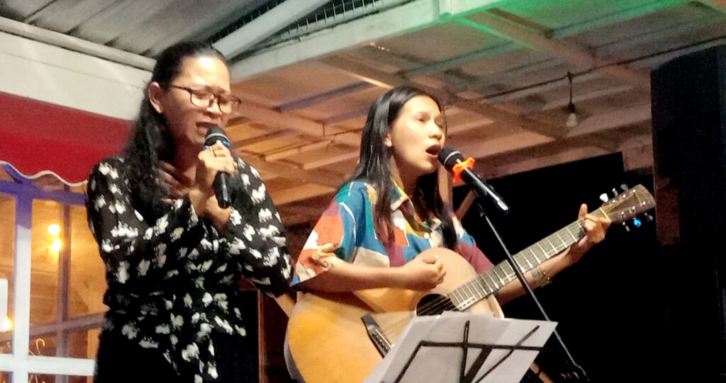 Two women perform on a small stage: one sings into a microphone with glasses, the other plays an acoustic guitar.