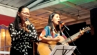 Two women perform on a small stage: one sings into a microphone with glasses, the other plays an acoustic guitar.