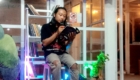 Person seated with a microphone reading from an open book inside a bookstore or cafe, bookshelves in the background and warm lighting.