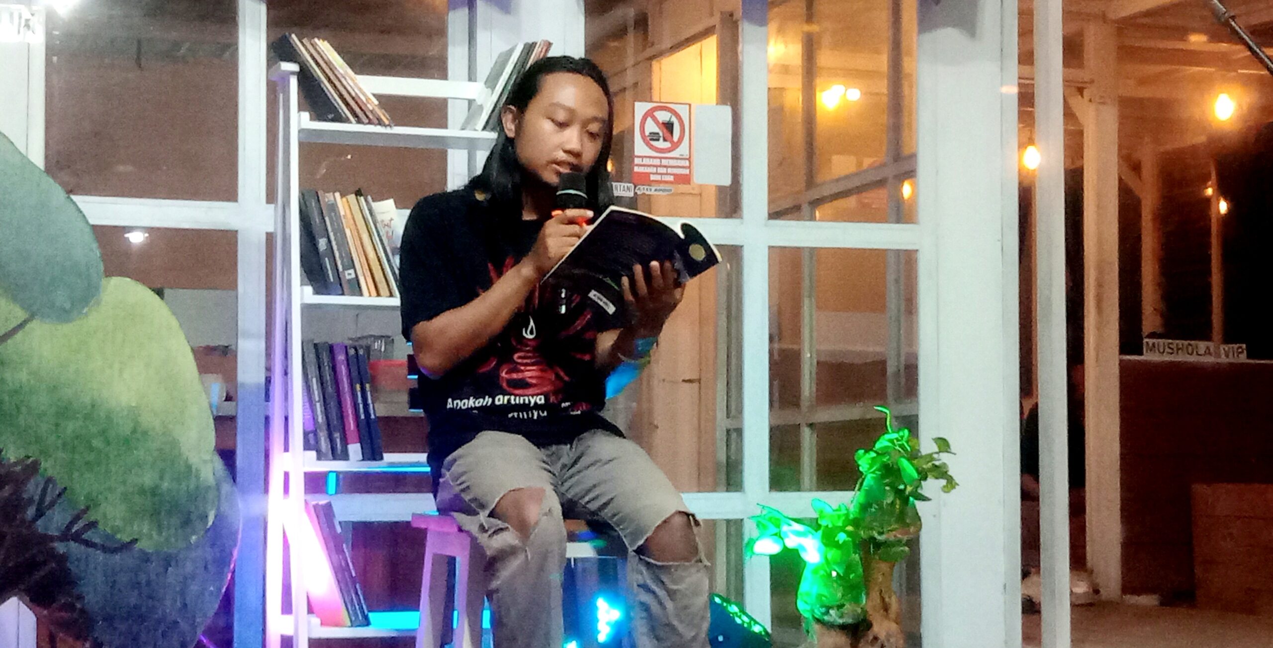 Person seated with a microphone reading from an open book inside a bookstore or cafe, bookshelves in the background and warm lighting.