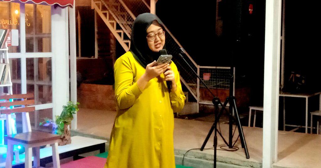 Woman in a bright yellow outfit and black hijab speaks into a handheld microphone at night on a stage.