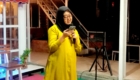 Woman in a bright yellow outfit and black hijab speaks into a handheld microphone at night on a stage.