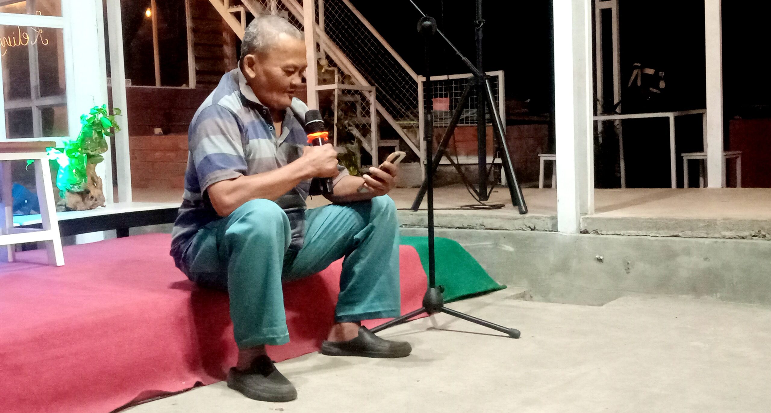 Man sitting on a red carpeted stage, holding a microphone in one hand and looking at his phone.