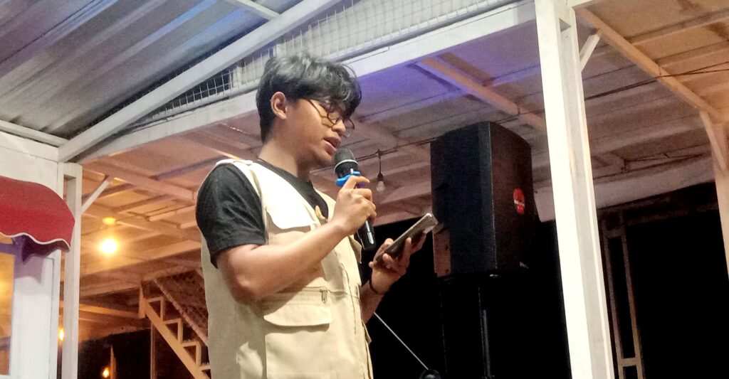 Young man wearing glasses and a beige vest speaks into a handheld microphone on a small stage, holding a phone or notes.