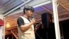 Young man wearing glasses and a beige vest speaks into a handheld microphone on a small stage, holding a phone or notes.