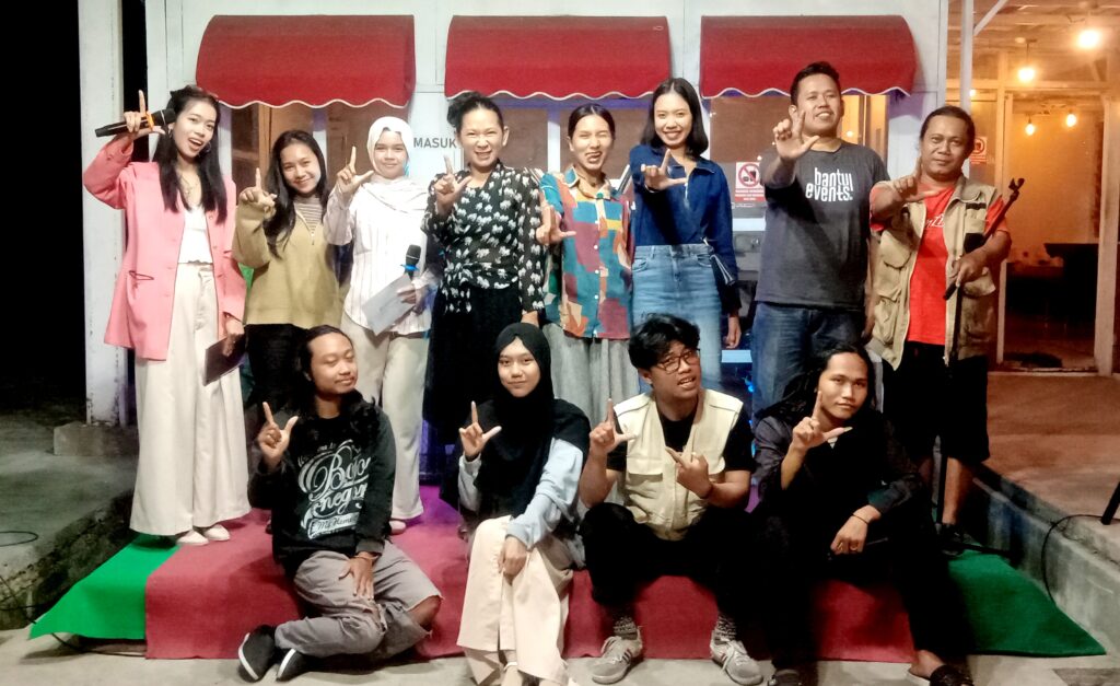 Group of young people posing on a stage with red awnings, making L-shaped hand gestures and smiling.