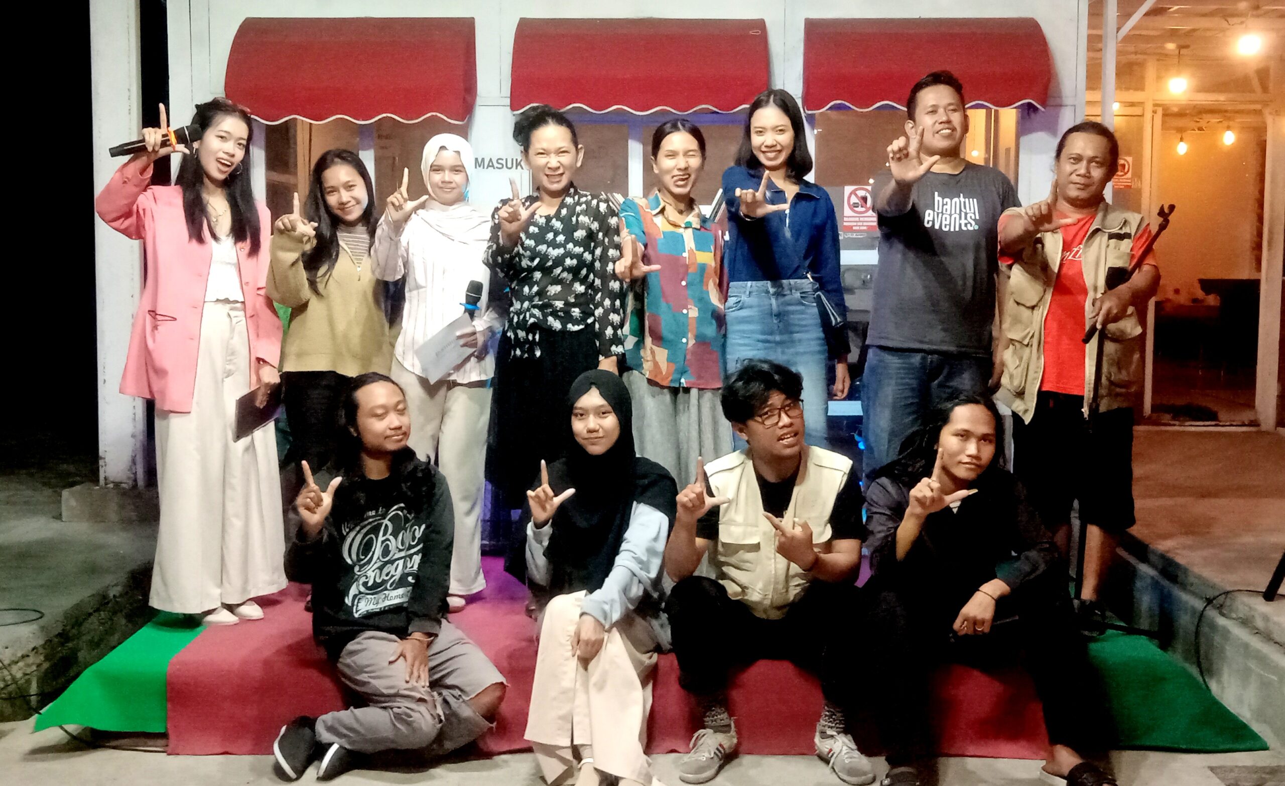 Group of young people posing on a stage with red awnings, making L-shaped hand gestures and smiling.