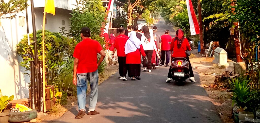 Group of people in red shirts walking down a residential street with greenery on both sides, a scooter parked on the right and a yellow banner in view.