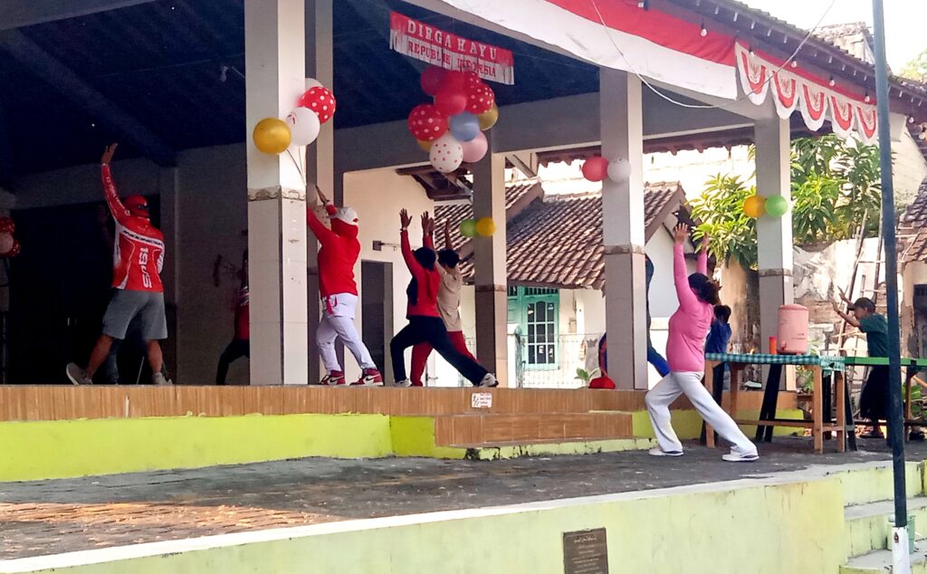 IMG_20260816_072927 - Mabur.co People dancing on an outdoor stage under a decorated pavilion with balloons and a red-white banner overhead.