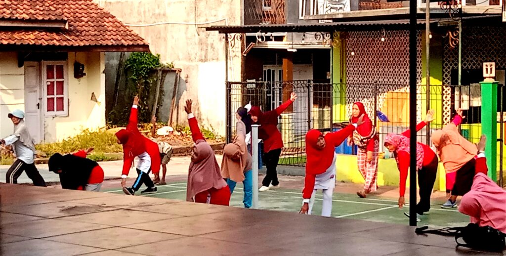 IMG_20260816_073003 - Mabur.co Group of people in hijabs and red outfits doing backward stretching on an outdoor court beside a colorful fence.