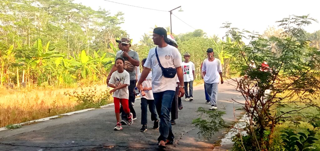 Group of people walking along a rural road bordered by banana trees and greenery on a sunny day.