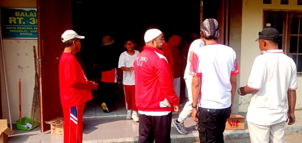 Group of people in red and white athletic outfits stand outside a doorway, with cleaning tools and boxes nearby in a sunny scene on a building entrance.