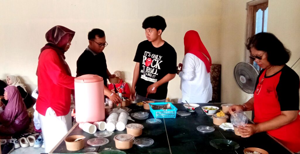 People in red and hijabs serve food and drinks at a long table, with a pink beverage dispenser and many disposable cups nearby.