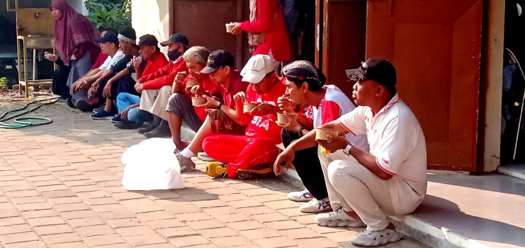 A row of people sitting on a curb outdoors, eating from brown cups, many wearing red shirts or caps.