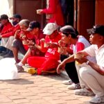 A row of people sitting on a curb outdoors, eating from brown cups, many wearing red shirts or caps.