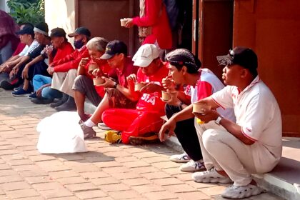 A row of people sitting on a curb outdoors, eating from brown cups, many wearing red shirts or caps.