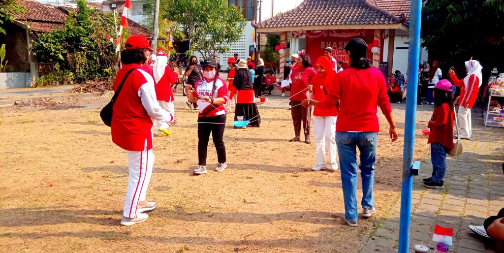 IMG_20260816_084733 - Mabur.co Outdoor community festival with people in red, playing a string-based balance game while others watch nearby.