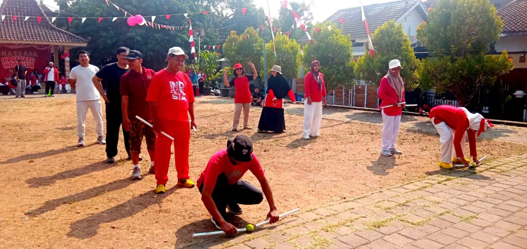 IMG_20260816_091101 - Mabur.co Group of people in red and white outfits playing a croquet-style game on a dusty ground while others watch at a festival, with bunting overhead.