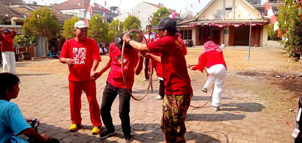 IMG_20260816_092815 - Mabur.co Group of people in red shirts playing with a looped rope in a sunny courtyard while others watch nearby at a festival.