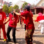 Group of people in red shirts playing with a looped rope in a sunny courtyard while others watch nearby at a festival.