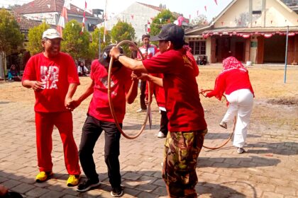 Group of people in red shirts playing with a looped rope in a sunny courtyard while others watch nearby at a festival.