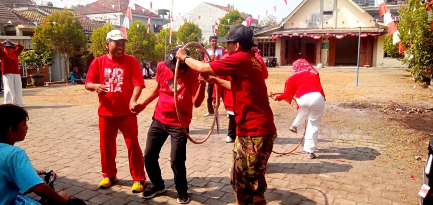 Group of people in red shirts playing with a looped rope in a sunny courtyard while others watch nearby at a festival.