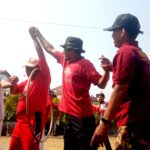 Group of people in red shirts dancing or celebrating outdoors at a festive event, with bunting flags and onlookers in the background.
