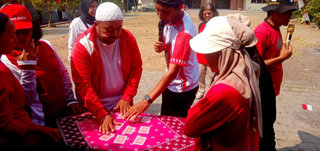 IMG_20260816_094734 - Mabur.co Group of people in red outfits gathered around a table with a pink patterned tablecloth, laying out playing cards outdoors.
