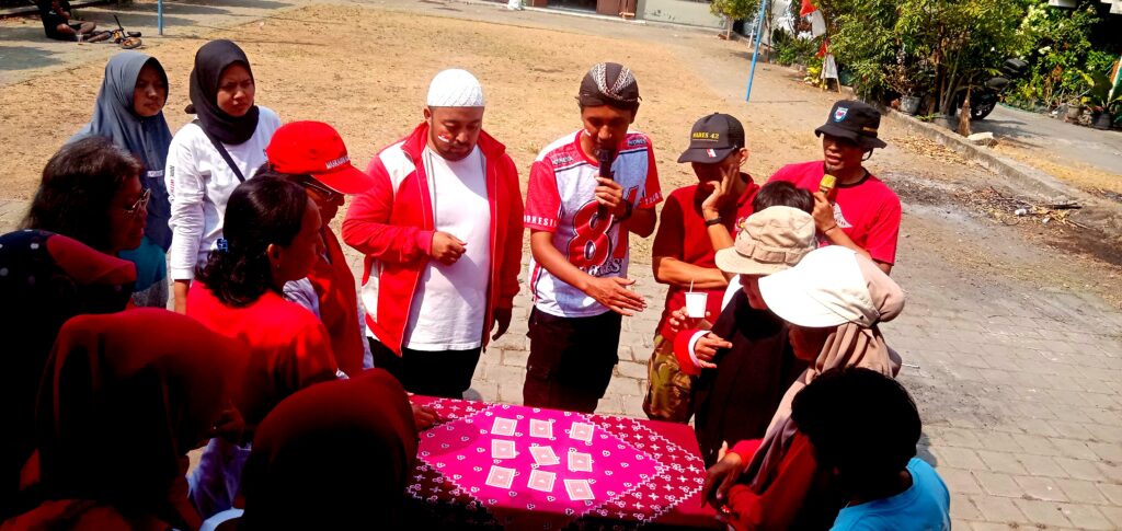 IMG_20260816_094935 - Mabur.co Group of people around a table with a pink card tablecloth outdoors; one man holds a microphone while others watch.