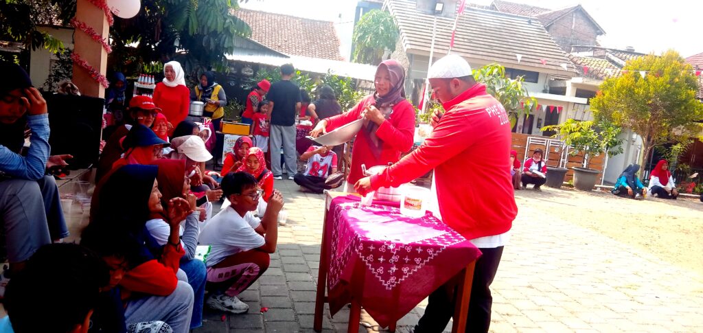 IMG_20260816_100644 - Mabur.co Group of people outdoors at a festive event; a man and woman in red shirts stand at a table handing out items to a seated crowd.